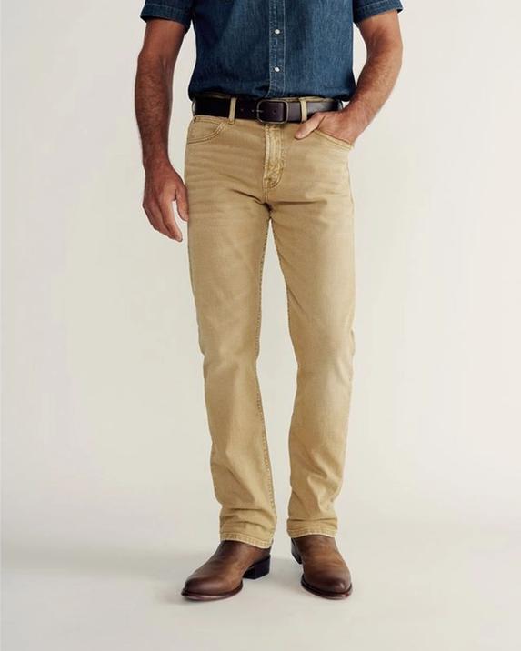 A person wearing a white t-shirt, olive green pants, and brown shoes stands with one hand in their pocket against a plain background.
