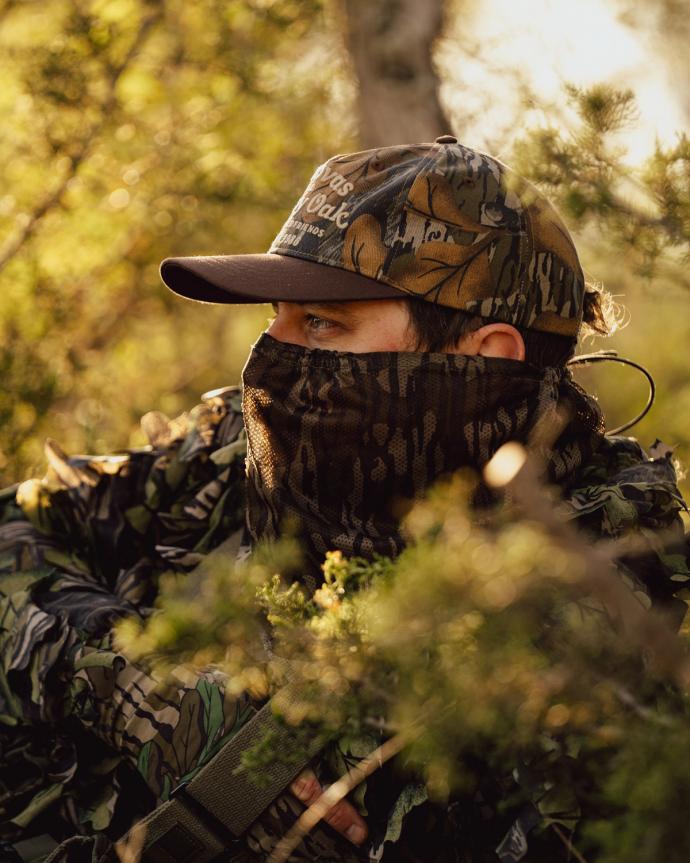Person in camouflage clothing and cap sits outdoors among foliage, with face partially covered by a camouflage mask, appearing to observe surroundings.