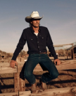A man in a white cowboy hat, dark shirt, and jeans sits on a wooden fence in an outdoor rural setting.
