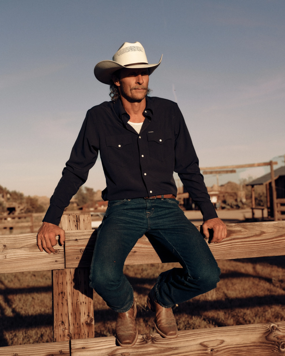 A man in a white cowboy hat, dark shirt, and jeans sits on a wooden fence in an outdoor rural setting.