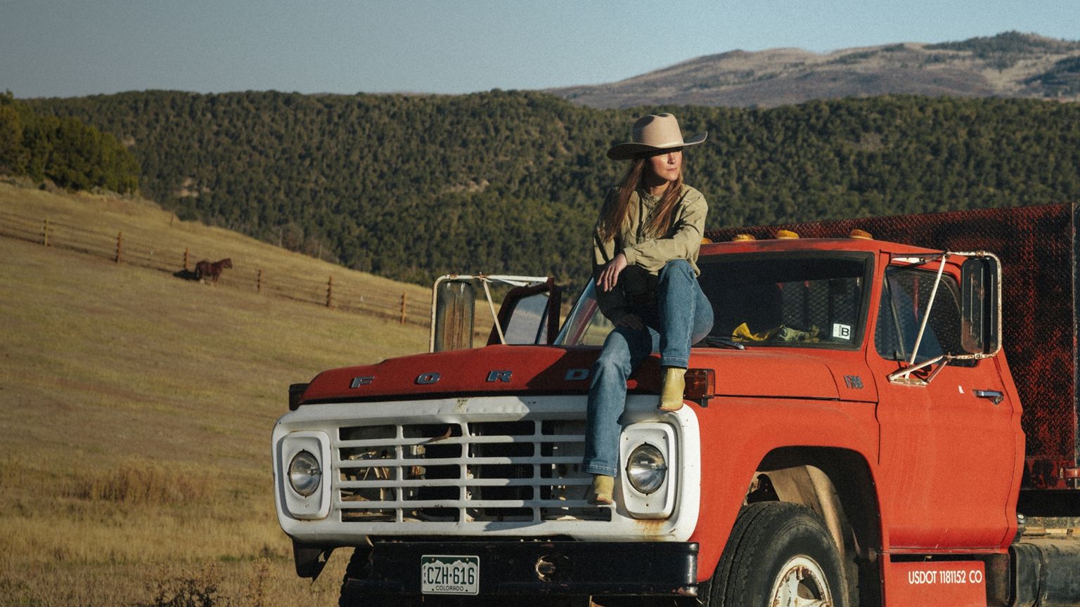 A woman in a cowboy hat sits on the hood of a red vintage Ford truck in a rural landscape with hills and a horse in the background.