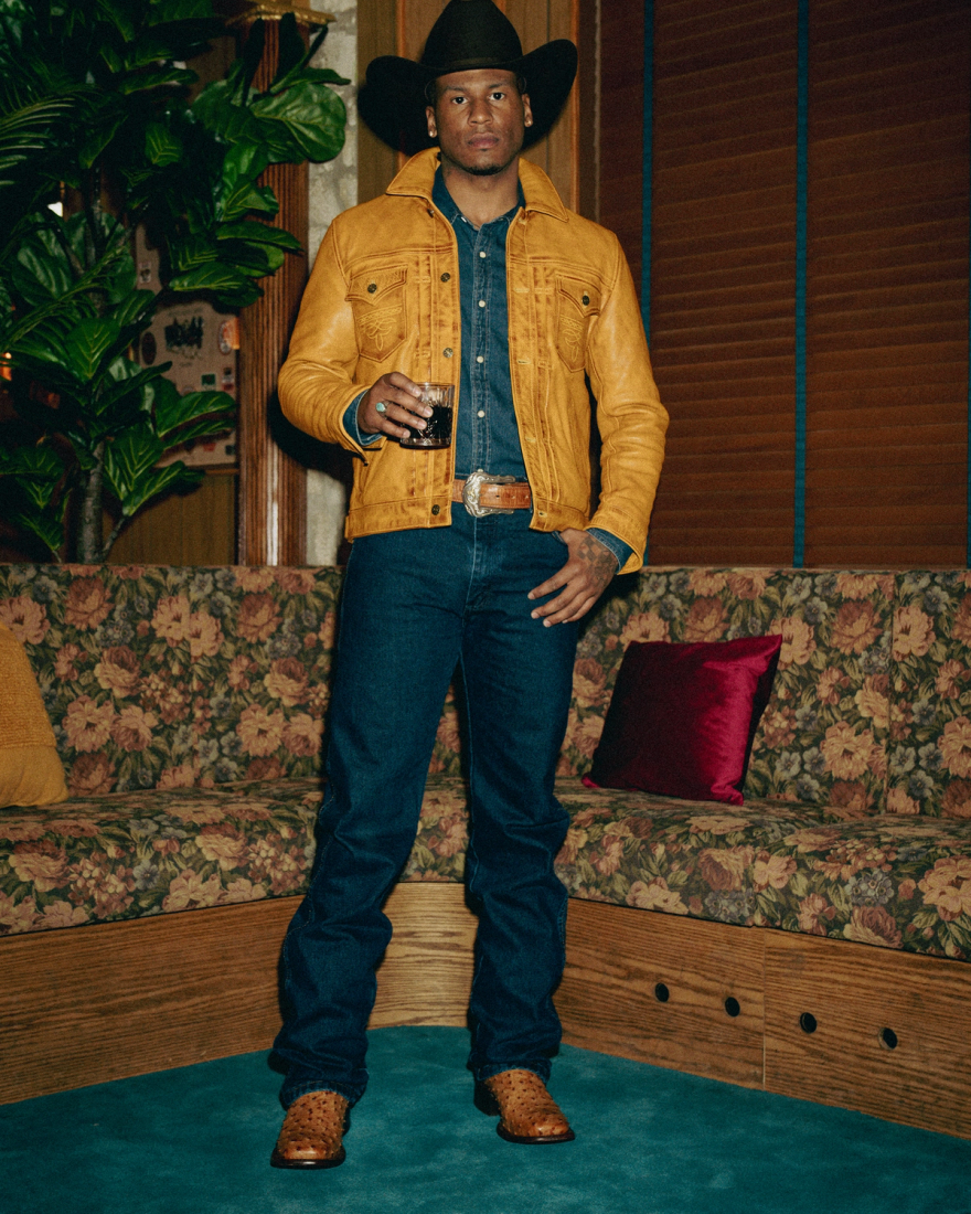 Person in a cowboy hat and denim outfit holds a drink, standing in a room with wooden panels, floral couch, and a large plant.