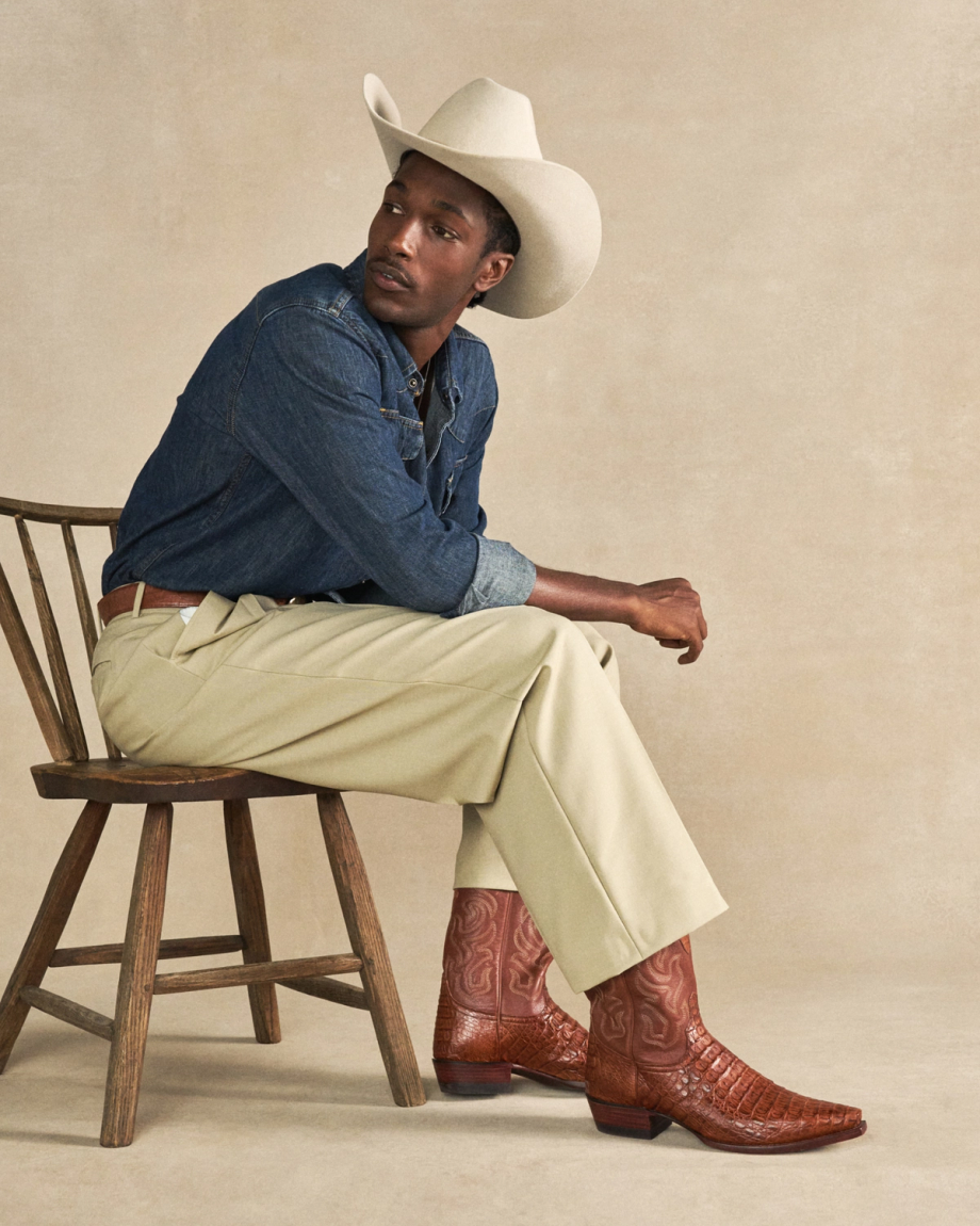 Man in a white cowboy hat, denim shirt, beige pants, and brown boots sits sideways on a wooden chair against a neutral background, sporting the rugged elegance of The Garrett Hornback Caiman boots in rich Pecan.