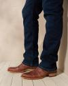 A person wearing blue jeans and brown leather shoes stands indoors on a light wooden floor against a beige wall.