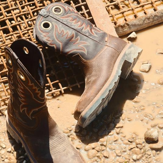 A pair of worn leather cowboy boots with decorative stitching sit on muddy ground beside a metal grate.