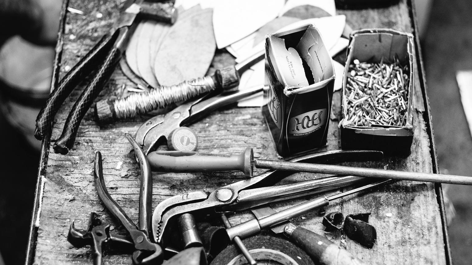 A cluttered workbench with various tools, including a hammer, pliers, wrenches, and boxes of nails. Black and white image.