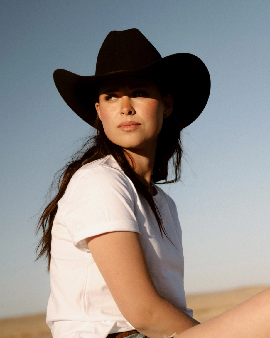 Woman in a white t-shirt and black cowboy hat sits outdoors, looking to the side, with a clear sky and desert landscape in the background.