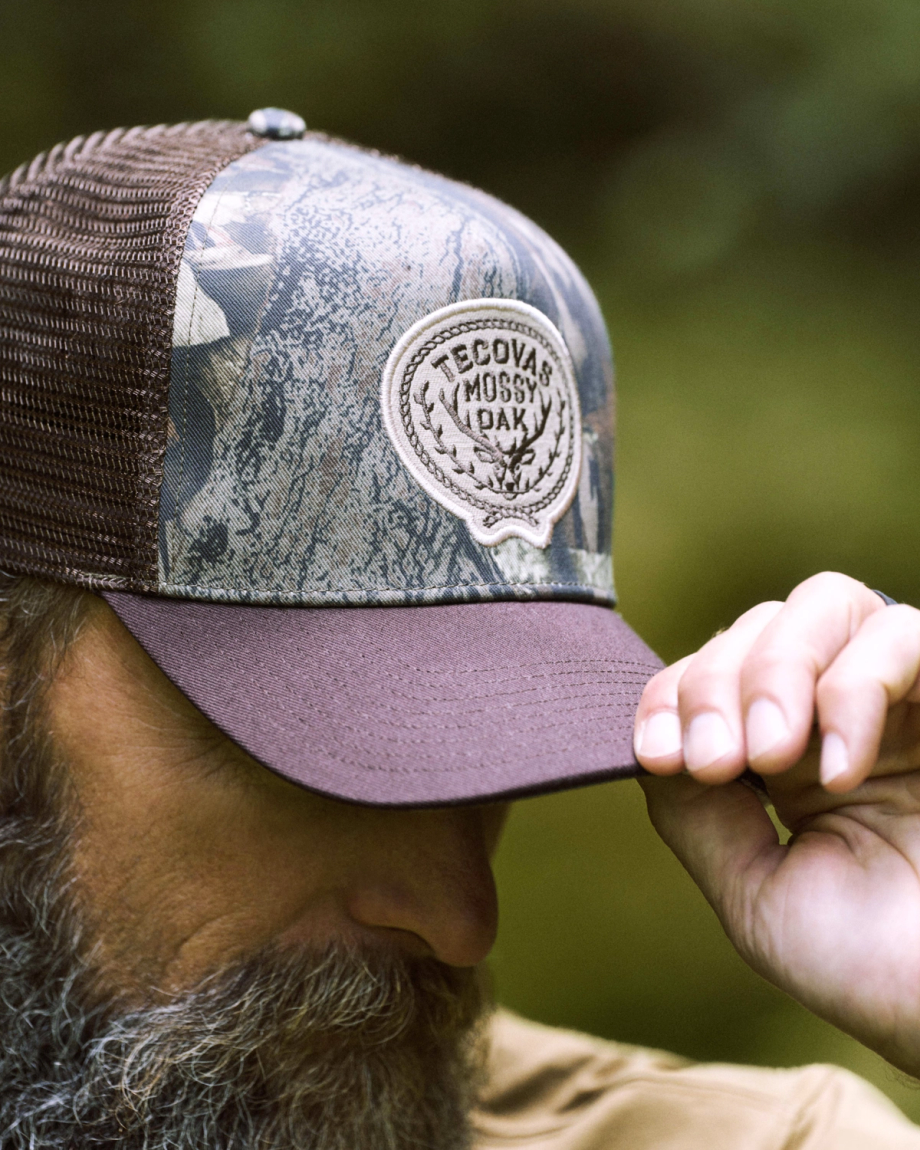 A person with a beard wears a camouflage mesh cap with a “Tecovas Mossy Oak” patch, holding the brim with their hand.