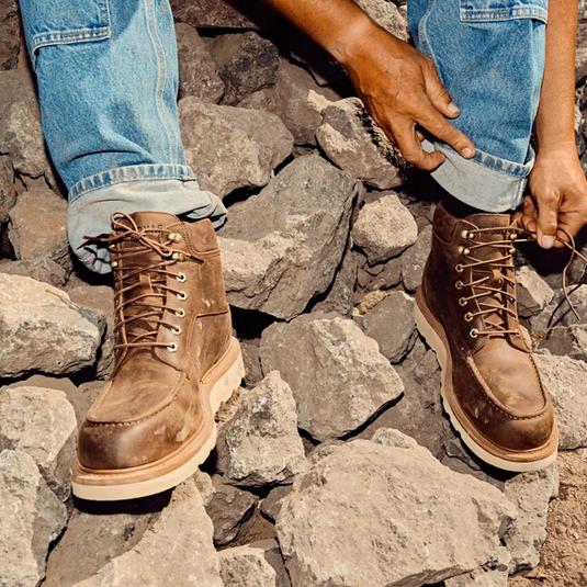 Person sitting on rocks, wearing blue jeans and brown lace-up boots, rolling up one pant leg with hands visible in the frame.