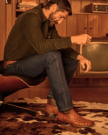 The Emmitt in Pecan Ostrich. A man with a beard sits on a modern chair in a wood-paneled room, wearing a dark shirt, blue jeans, and Pecan Ostrich boots, with a vintage TV and record player in the background.
