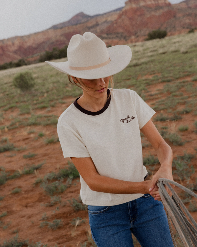 Person in a wide-brimmed hat and "Good Luck" shirt standing outdoors, holding a net. Desert landscape with red rocks and sparse vegetation in the background.