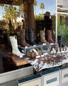 A store window display featuring various cowboy boots in multiple colors and patterns, belts, and leather accessories set on a table with a cowhide covering. Palm trees and reflections are in the background.