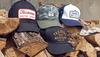Four caps with various designs, including logos and text, are displayed on a stack of firewood against a wall.