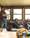 Three men in camouflage clothing relax and chat in a cozy room with large windows, holding drinks.