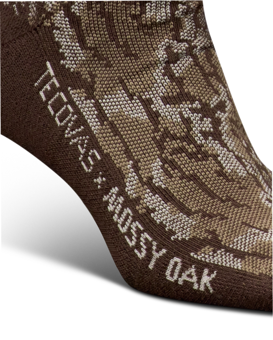 Close-up of a brown sock with a camouflage pattern and the text "TECOVAS x MOSSY OAK" knitted into the fabric near the ankle.