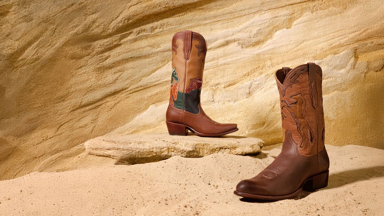 Two brown cowboy boots with decorative stitching are displayed on sand and rock against a textured, tan stone background.
