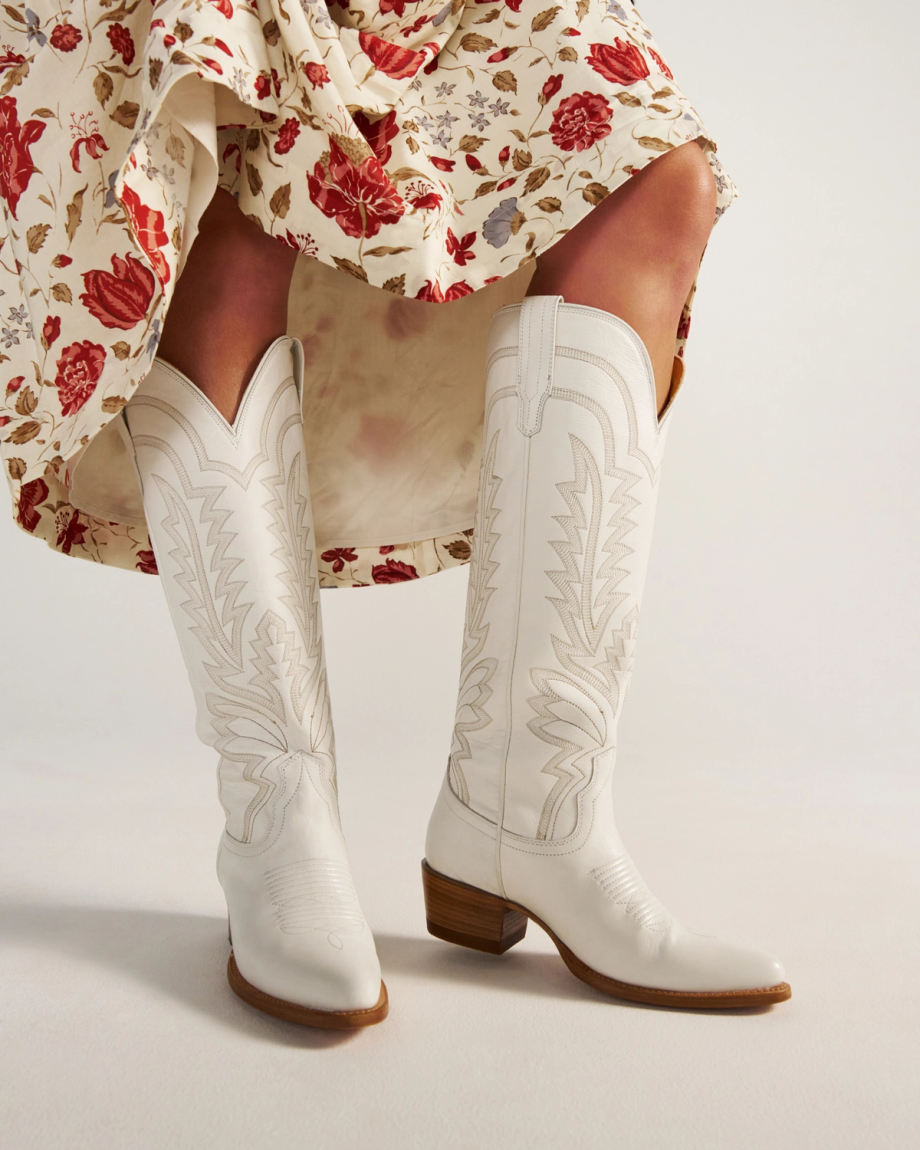 The Abby Wide Calf in Snow Cowhide. Person wearing a floral skirt and white Snow Cowhide embroidered cowboy boots with a wooden heel, standing on a light background.
