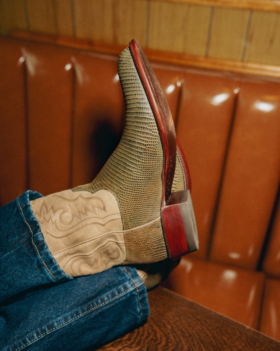 Close-up of a person's feet wearing textured cowboy boots with patterned shafts, resting on a brown cushioned seat. The person is dressed in blue jeans.