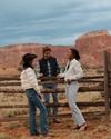 Three people, dressed in casual and cowboy-style clothing, are standing and talking near a wooden fence in a desert landscape with rocky formations in the background.