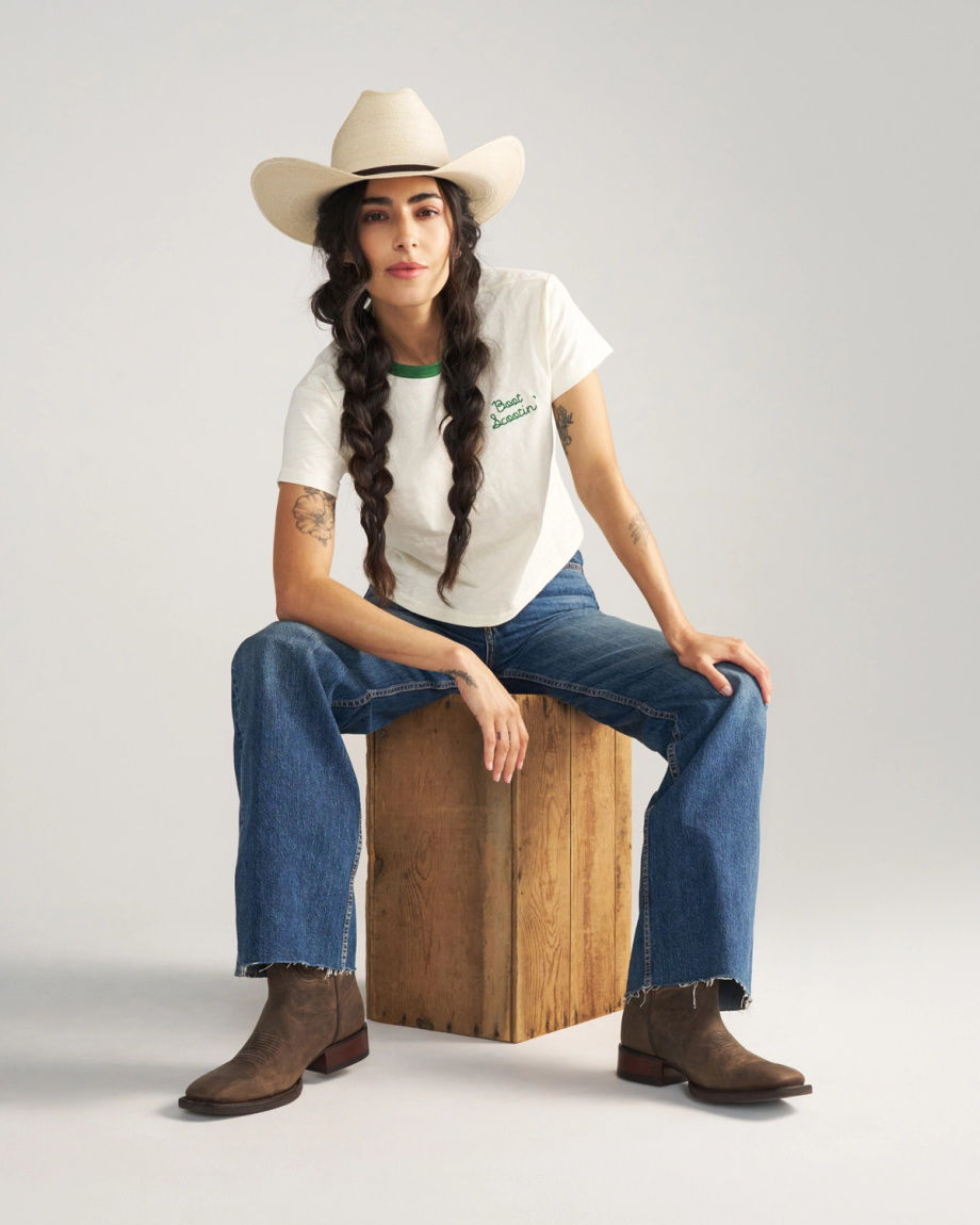 The Tess in Cafe Goat. A person wearing a cowboy hat, white t-shirt, blue jeans, and brown boots sits on a wooden box against a plain background, evoking the relaxed charm of Cafe Goat.