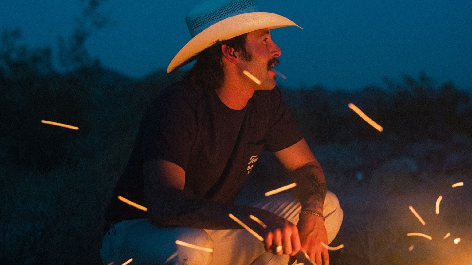 A man in a white cowboy hat and dark shirt sits by a campfire at dusk, surrounded by flying embers.