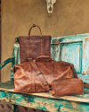 A collection of brown leather bags, including a tote, duffel, and small pouch, are displayed on a worn, turquoise wooden bench outdoors against a stucco wall with a mounted animal skull above.