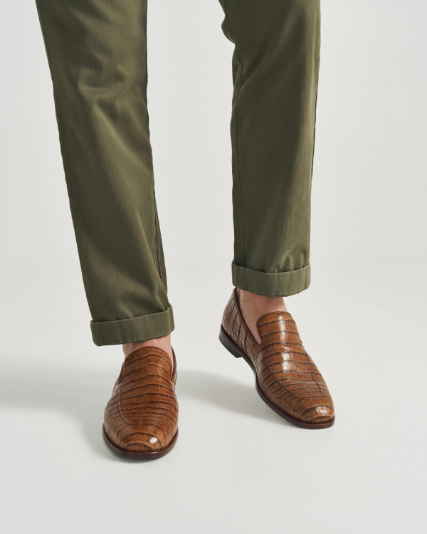 The Bennett in Saddle Alligator. A person wearing olive green pants and The Bennett Alligator brown crocodile-patterned leather loafers stands on a white background.
