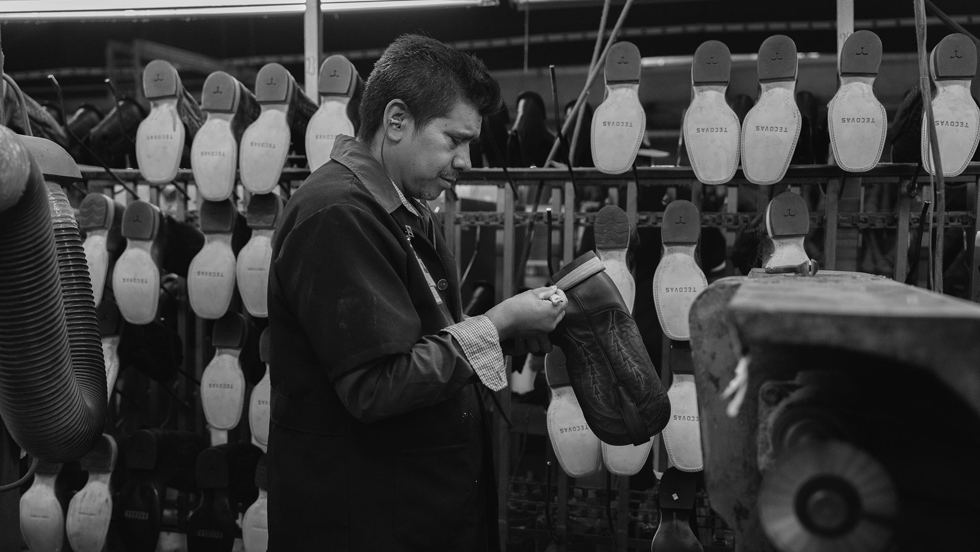A worker inspects a shoe sole in a factory, with multiple shoe soles hanging on racks in the background.