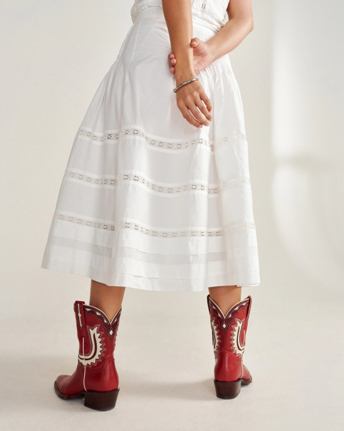 Person wearing a white skirt and red cowboy boots, standing with hands clasped behind their back.