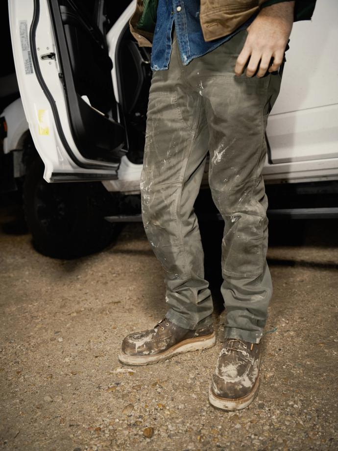 A person standing next to an open vehicle door, wearing a denim shirt, khaki pants, and work boots covered in mud.