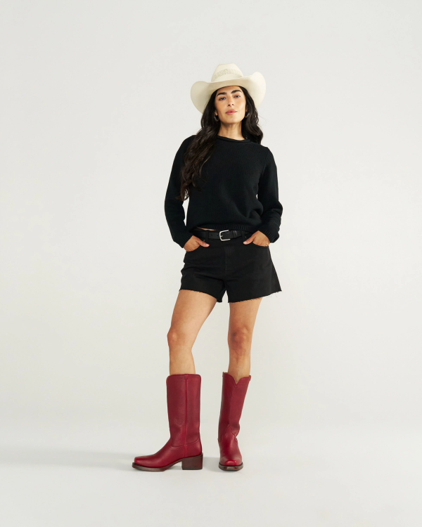 The Charlie in Scarlet Cowhide. Woman in a white cowboy hat, black sweater, black shorts, and striking Scarlet Cowhide red cowboy boots stands against a plain white background.