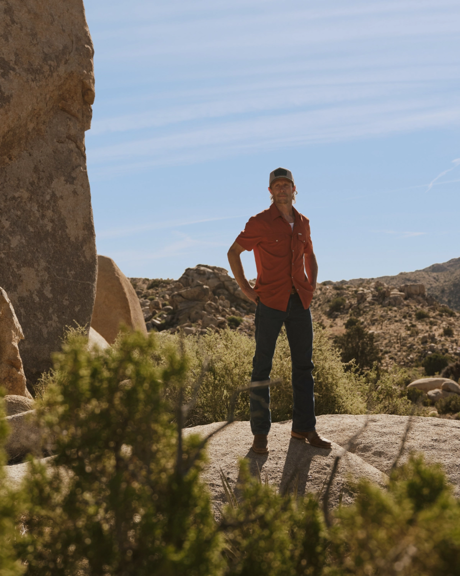 A man in a red shirt and jeans stands on a rocky landscape with desert vegetation and large boulders under a blue sky.
