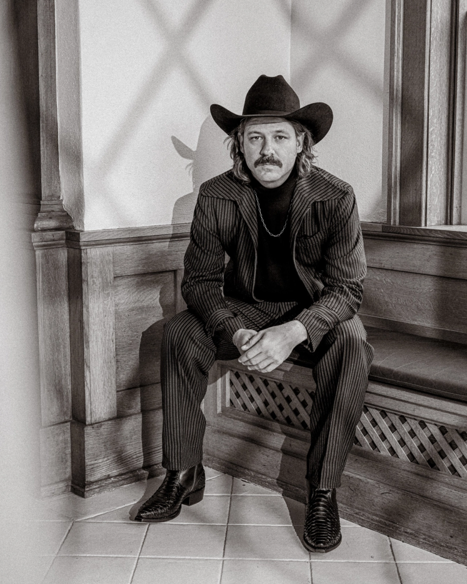 A person in a pinstripe suit and cowboy hat sits on a bench. The setting includes wooden paneling and tiled flooring. The image is black and white.