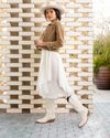 A woman in a white dress, brown jacket, and white cowboy boots stands confidently in front of a geometric wooden wall, wearing a wide-brimmed hat.