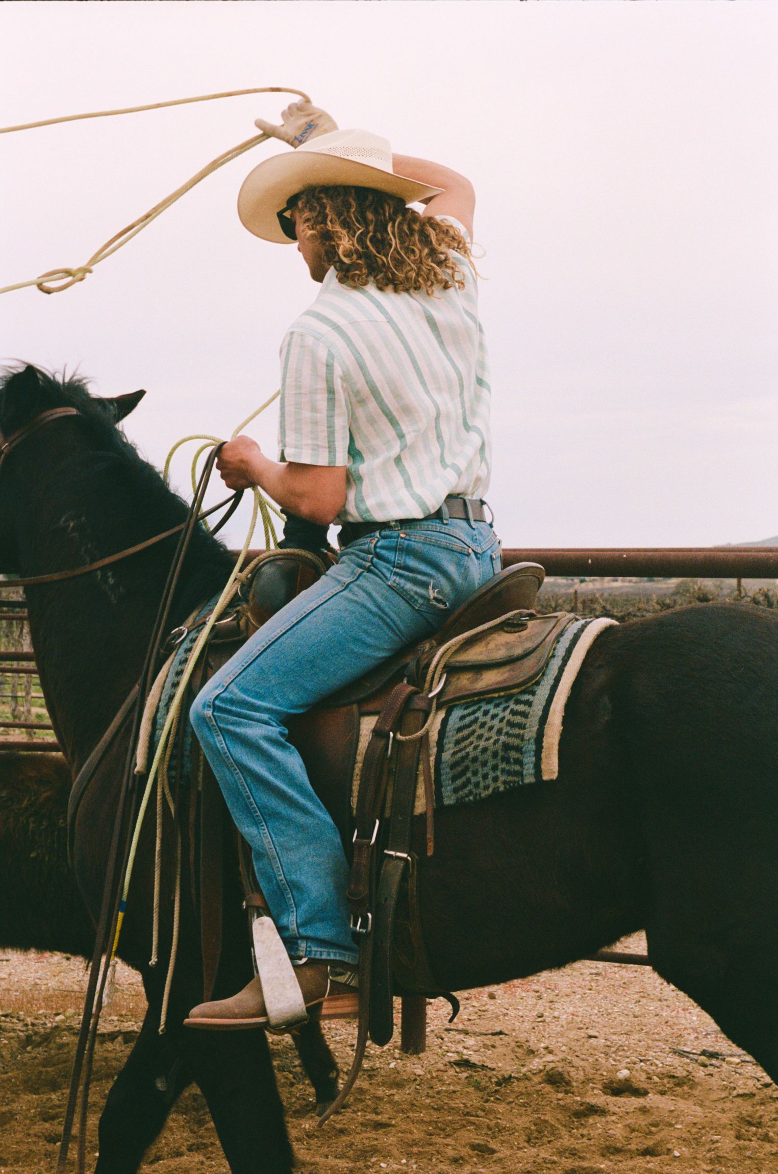 A person in a striped shirt, jeans, and cowboy hat rides a saddled horse, holding a lasso above their head in a ranch setting.