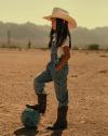 A child wearing a cowboy hat, boots, and overalls stands on a dirt field with one foot on a blue soccer ball, mountains visible in the background.