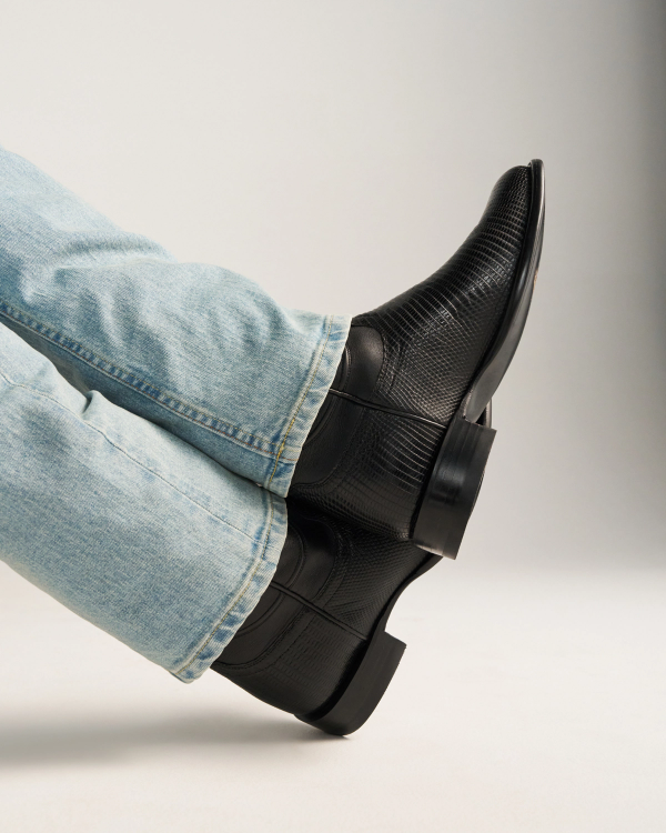 The Earl in Obsidian Lizard. A person wearing light blue jeans and The Earl Lizard black textured leather dress boots is sitting with their legs crossed against a neutral background.