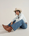 Person sitting on the floor wearing a cowboy hat, light blue shirt, jeans, and brown cowboy boots against a plain background.