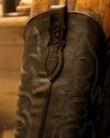 Close-up of a dark leather cowboy boot with decorative stitching and embossed detailing, leaning against a wooden surface.
