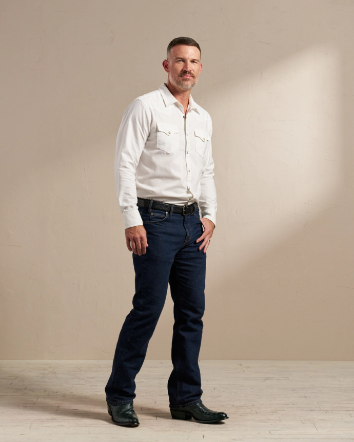 A man stands against a plain beige background wearing a white button-up shirt, dark blue jeans, a belt, and black boots.