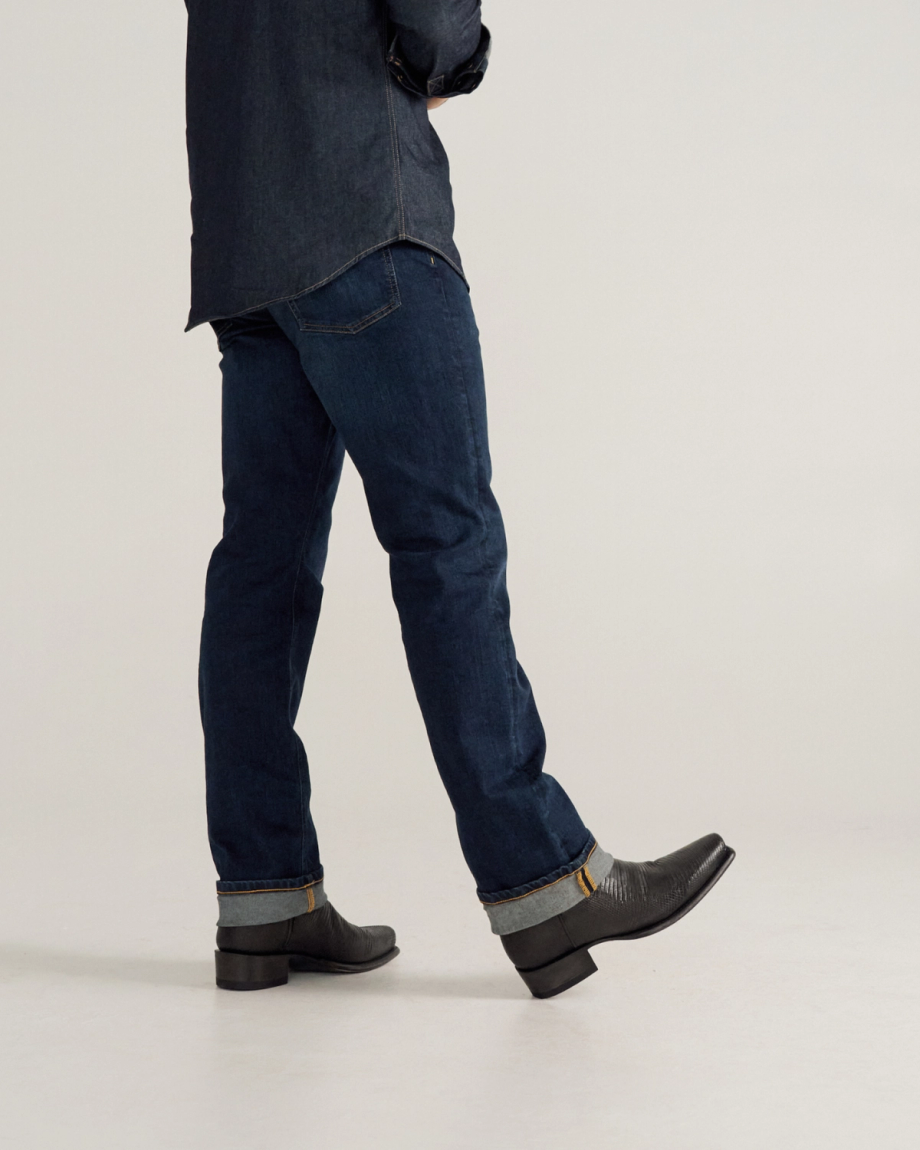 Person wearing dark blue jeans, a dark shirt, and black boots is walking, shown from the shoulders down against a plain background.