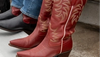 Close-up of red cowboy boots with intricate stitching, worn by someone standing on a textured surface. Another person wearing black and red cowboy boots is partly visible beside them.