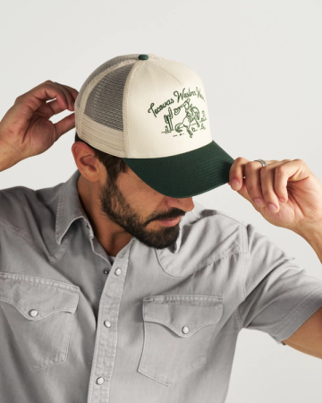 Man wearing a grey button-up shirt and adjusting a beige and green trucker hat with a western-themed illustration and text.