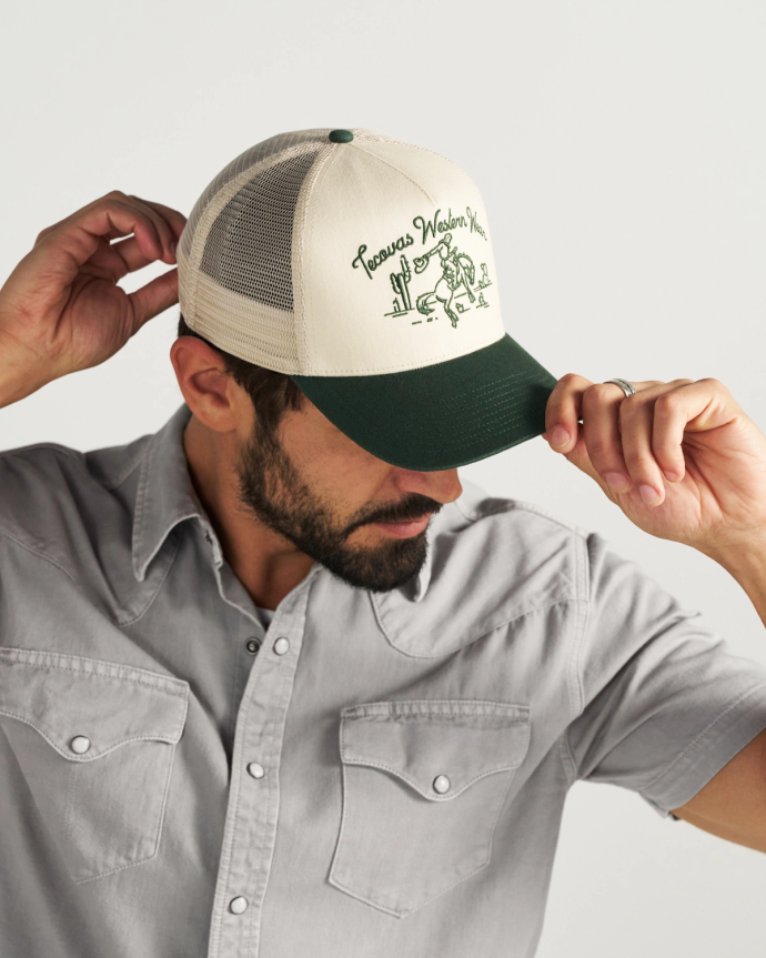 Man wearing a grey button-up shirt and adjusting a beige and green trucker hat with a western-themed illustration and text.