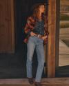 A woman in a cowboy hat, patterned jacket, denim shirt, and jeans stands in a doorway, looking to the side with hands in her pockets.