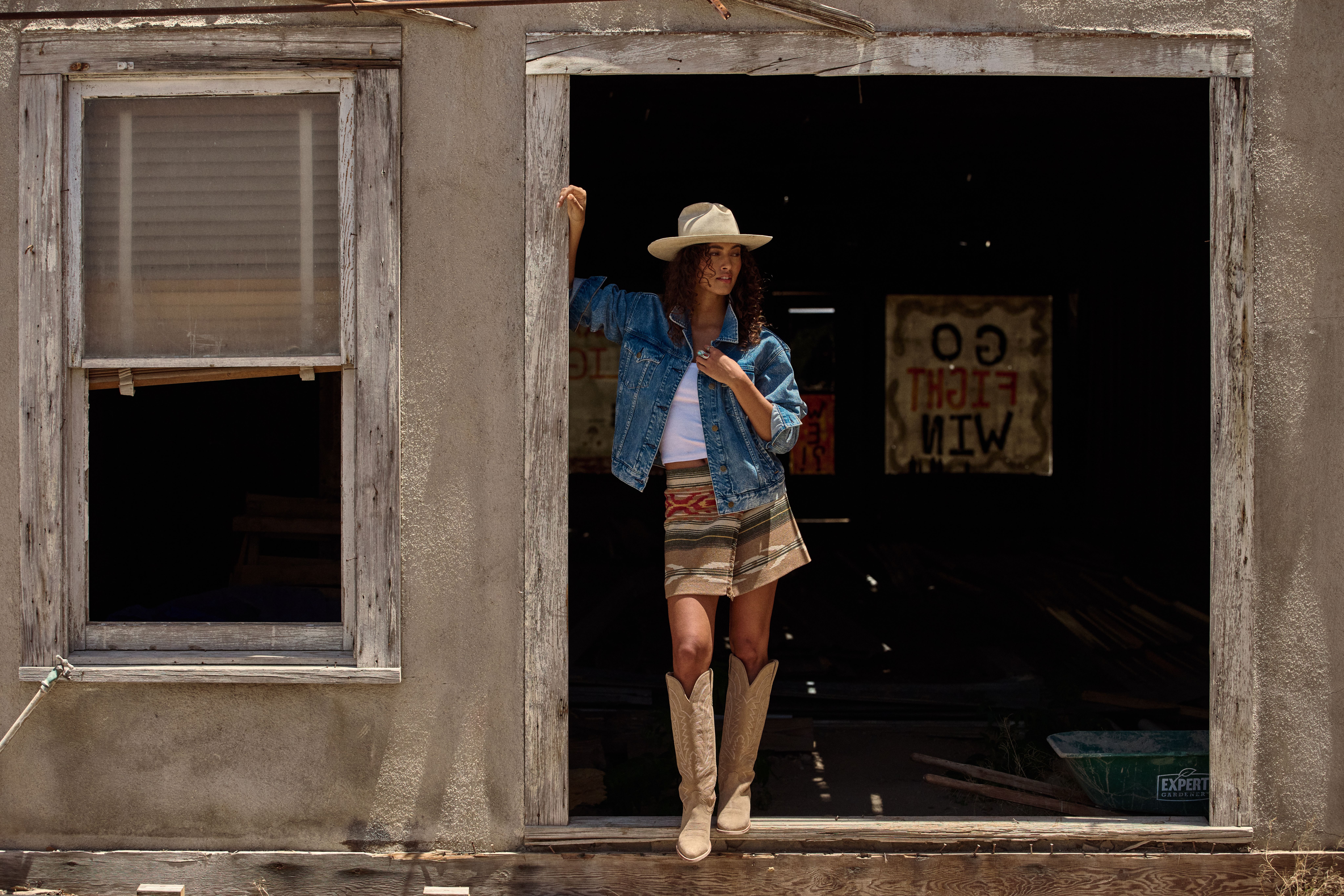 A woman in a hat, denim jacket, patterned skirt, and boots stands in the doorway of a rustic building with faded wooden frames.