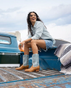 A person with curly hair, wearing a light denim shirt, denim shorts, and boots, sits on the edge of a blue vehicle's cargo area, smiling and looking to the side.