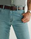 A person wearing light blue jeans, a dark brown belt, a grey shirt, and a wristwatch with their right hand in their pocket.
