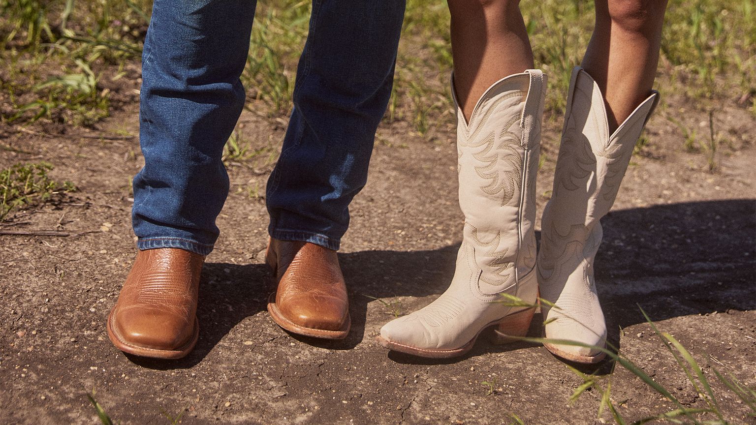 A pair of people standing on a dirt path, one wearing brown leather boots and blue jeans, and the other wearing white cowboy boots and a skirt.