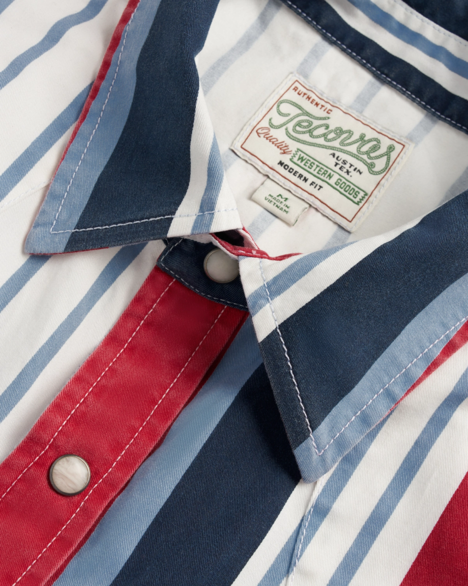 Close-up of a collared shirt with vertical red, white, and blue stripes, snap buttons, and a sewn label that reads "Genuine Western Goods, Modern Fit.